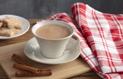 Fotografia em tons de vermelho e marrom de uma bancada, ao centro uma xícara com chocolate quente saindo fumaça e um pires abaixo. Ao lado pedaços de canela, biscoitinhos e uma pano xadrez vermelho e branco.